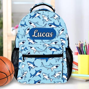 May include: A light blue backpack with a repeating pattern of cartoon sharks. The backpack has black mesh side pockets and a black handle. The name "Lucas" is displayed on a blue oval patch on the front.