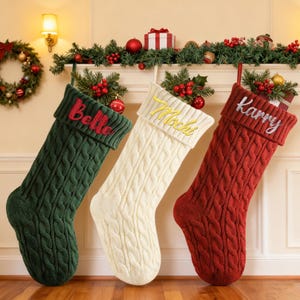 May include: Three cable-knit Christmas stockings in green, cream, and red hang from a mantel. Each stocking has a folded cuff with a name embroidered in a contrasting color: "Belle" in red, "Mochi" in yellow, and "Karry" in silver.