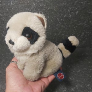 Op de afbeelding: Een pluche wasbeer speelgoed met een crèmekleurig lichaam, zwart masker rond de ogen en een gestreepte staart. Het speelgoed heeft een klein, rond, rood en blauw label. De wasbeer zit in de hand van een persoon.