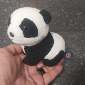 Op de afbeelding: Een kleine, zachte panda beer in zwart en wit. De panda heeft zwarte vlekken rond de ogen, oren en op het lichaam. Een klein blauw label met een rode cirkel is aan de zijkant van de panda bevestigd.