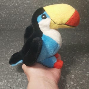 Puede incluir: Un juguete de peluche tucán con cabeza negra, cuerpo azul, pico amarillo y detalles rojos. El tucán está sentado en una mano.