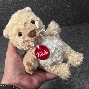May include: A small plush animal toy with cream-coloured fur, a white belly, and dark brown accents on the nose and paws. The toy features black eyes, white thread whiskers, and a round red tag around its neck with the white text Trudi printed on it.