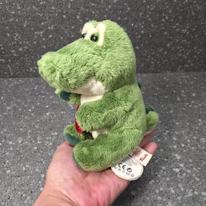 Op de afbeelding: Een kleine, groene knuffelkrokodil met een crèmekleurige buik, grote gele en zwarte ogen en een klein rood hartvormig detail op de borst. Het knuffeldier is gemaakt van zachte, gestructureerde stof en heeft een wit merklabel aan de zijkant.