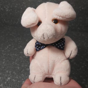 Op de afbeelding: Een roze pluche varken met een donkerblauwe strik met witte stippen. Het varken heeft een kleine snuit en grote oren.