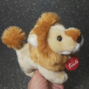 Op de afbeelding: Een pluche leeuwenknuffel met een bruine manen en een beige lichaam. Het speelgoed heeft een rood label met het woord "Trudi" erop.