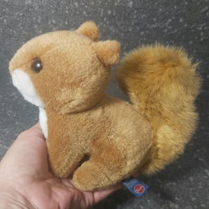 Op de afbeelding: Een pluchen eekhoorn met een pluizige, lichtbruine staart en zacht, bruin haar. De eekhoorn heeft een witte borst en een klein, rond, donker oog. Het speelgoed wordt in een hand gehouden en een blauw label met een rode cirkel is zichtbaar.