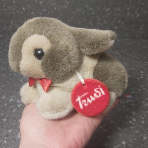 Op de afbeelding: Een klein pluche konijn in grijs en beige met een rode strik. Het konijn heeft een rood label met de tekst "Trudi" eraan.