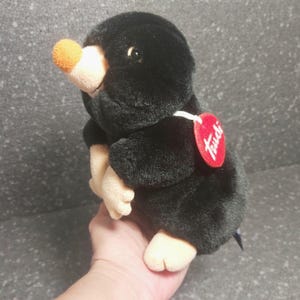 Op de afbeelding: Een zwart pluche mol speelgoed met een bruine snuit en poten. De mol heeft een kleine, oranje neus en een rood hartvormig label met het woord "Trudi". Het speelgoed is zacht en knuffelig.