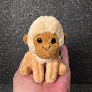 May include: A small plush monkey toy with light cream-colored fur on its head and chest, and tan-colored fur on its face and limbs. The stuffed animal features two round, black plastic eyes and a simple stitched brown V-shaped mouth.