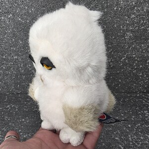 Op de afbeelding: Een witte uil knuffel met gele ogen en zwarte accenten. De uil heeft een zachte, donzige textuur en een klein label. Het speelgoed is ontworpen om op een sneeuwuil te lijken.