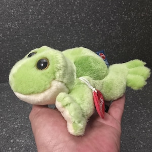 Op de afbeelding: Een groene en crèmekleurige pluche kikker. De kikker heeft grote, zwarte ogen en een glimlachende uitdrukking. Een rood label is aan het speelgoed bevestigd. Het is een knuffel.