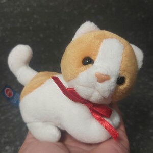Op de afbeelding: Een zacht speelgoedkatje met een witte basis en oranje vlekken op het hoofd en de rug. Het heeft zwarte ogen, een roze neus en een rode strik. De kleine, knuffelige kat is ontworpen voor kinderen.