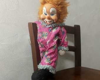 May include: A vintage clown doll with a smiling face, orange hair, and a pink floral patterned outfit. The doll is seated on a wooden chair, with one black shoe visible. The clown's outfit has ruffled trim.