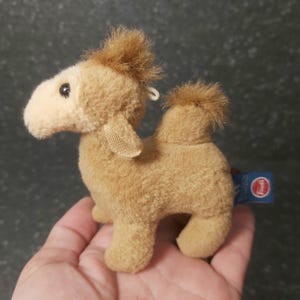 Op de afbeelding: Een klein, pluche kameel speelgoed in tinten bruin en beige. De kameel heeft een pluk bruin haar op zijn hoofd en een klein label. Het speelgoed wordt in een hand gehouden.