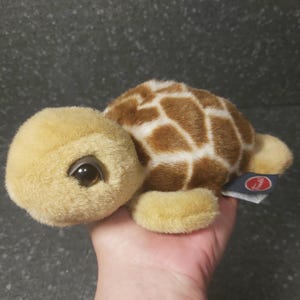 Op de afbeelding: Een pluche schildpad met een beige kop en ledematen, en een bruin-wit patroon op de schild. De schildpad heeft grote, donkere ogen en een klein label aan de zijkant. Het speelgoed is zacht en knuffelbaar.