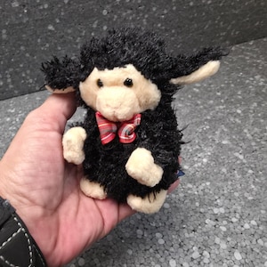 Op de afbeelding: Een kleine pluche knuffel in de vorm van een lammetje met zwarte vacht op het hoofd en lichaam, een crèmekleurig gezicht, oren en pootjes, en een rood, groen en wit geruite vlinderdas om de nek. Het speelgoed heeft donkere ogen en een zachte textuur.