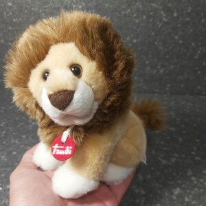 Puede incluir: Un león de peluche marrón y beige con la cara blanca y una etiqueta roja que dice "Trudi" en el pecho.