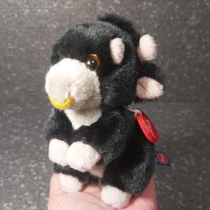 Puede incluir: Vaca de peluche negra y blanca con un anillo de nariz amarillo. El juguete tiene una etiqueta roja adjunta.