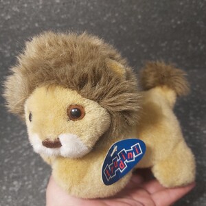 Op de afbeelding: Een pluchen leeuwenspeelgoed met een bruin lichaam en een bruine manen en staart. Het speelgoed heeft een label met de tekst "Trudini" in blauw en wit. De leeuw heeft bruine ogen en een bruine neus.