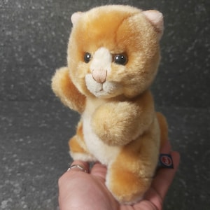Puede incluir: Un peluche de gato atigrado naranja con pecho y patas blancas. El peluche tiene ojos negros, nariz rosa y una pequeña etiqueta azul. El gato está sentado con una pata levantada.