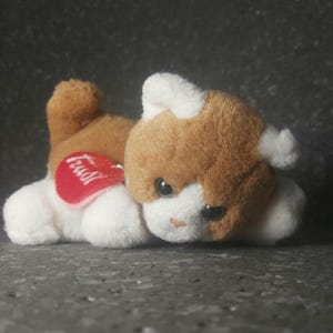 Puede incluir: Un gato de peluche marrón y blanco, con una etiqueta roja en forma de corazón. El gato tiene ojos grandes y expresivos y está tumbado. La etiqueta dice "Trudi".