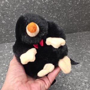 May include: A plush mole toy featuring soft black fur, a round orange nose, and cream-coloured paws. The stuffed animal has a small red bow tie detail at the neck. The toy is compact and rounded in shape, with visible stitching and soft textures.