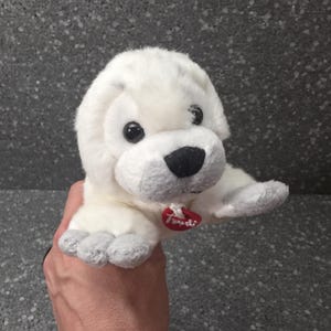Op de afbeelding: Een witte pluche puppy met zwarte ogen en een zwarte neus. De puppy heeft een rood hartvormig label. De puppy ligt met zijn poten uitgestrekt.