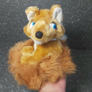 Puede incluir: Un juguete de peluche de zorro con pelaje naranja y blanco, ojos azules y una nariz negra. El zorro está sentado con su cola esponjosa enrollada alrededor de su cuerpo. El juguete está hecho de material suave y afelpado.