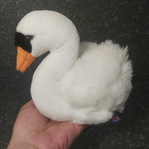 Op de afbeelding: Een zacht, wit zwaan speelgoed met een zwart masker rond de ogen en een oranje snavel. De zwaan is gemaakt van zacht, pluizig materiaal en is ontworpen om op een echte zwaan te lijken. Het speelgoed wordt in een hand gehouden.