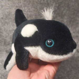 Op de afbeelding: Zwarte en witte pluche orka met een witte vlek op zijn rug en een blauw oog. De orka heeft een witte buik en een zwarte rugvin.