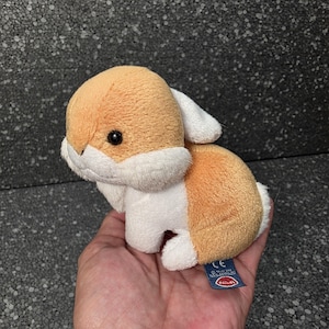 Op de afbeelding: Een kleine pluche konijnknuffel met zachte beige en witte vacht, zwarte plastic ogen en een klein blauw stoffen label aan de zijkant. Het knuffeldier heeft een ronde kop, hangende oren en een compact lichaam met een witte borst en pootjes.