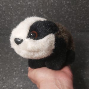 Op de afbeelding: Een pluche speelgoed das met een zwart en wit gezicht, een zwart lichaam en bruine vacht op de rug. De das heeft een zwarte neus en bruine ogen. Een zacht en knuffelbaar speelgoed voor kinderen of verzamelaars.