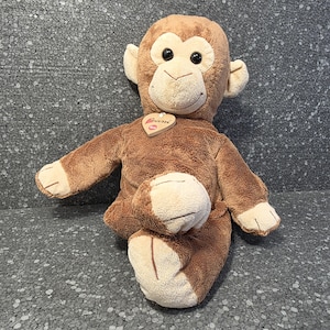 Op de afbeelding: Een bruine pluchen aap met een lachend gezicht, grote oren en een hartvormig label. Het knuffelbeest zit met uitgestrekte armen en benen. Het label leest "Cousi".
