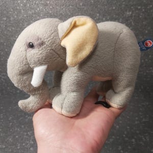 Puede incluir: Un juguete de peluche de elefante gris con una oreja amarilla y un colmillo blanco. El juguete está sentado en la mano de una persona.