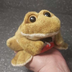 Op de afbeelding: Een pluche bruine kikker speelgoed met grote zwarte ogen en een rode mond. De kikker houdt een klein rood voorwerp in zijn mond.