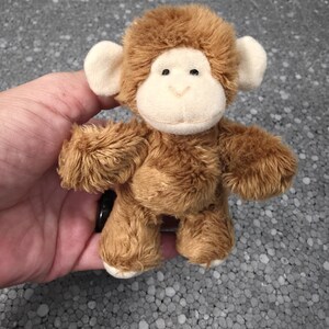 May include: A small plush monkey toy featuring soft brown fur, a cream-colored face, and rounded cream-colored ears. The stuffed animal has black bead eyes and a stitched nose and mouth, with a fuzzy texture covering its body and limbs.