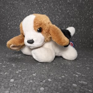 Op de afbeelding: Een pluche hondenspeeltje met een driekleurige vacht van wit, lichtbruin en zwart bont. Het knuffeldier heeft lange, hangende oren, een witte bles op het voorhoofd, een zwarte neus en donkerbruine ogen, afgebeeld in een liggende positie.