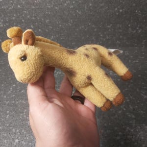 Op de afbeelding: Een pluche giraf in tinten bruin en beige. De giraf heeft bruine vlekken, oren en hoeven. Het speelgoed is zacht en knuffelbaar, met een klein label eraan.
