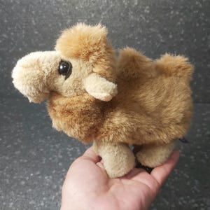 Op de afbeelding: Een klein, bruin pluche kameel speelgoed met zwarte ogen. De kameel staat op de hand van een persoon.