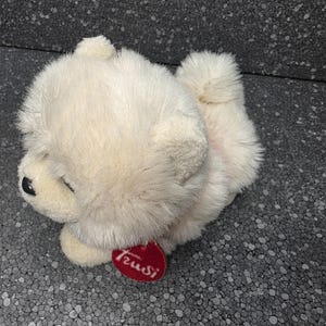 Op de afbeelding: Een pluche hond met crèmekleurige, pluizige vacht, kleine ronde oren en een zwarte neus. Een klein, hartvormig rood label met de witte tekst Trudi is bevestigd aan de nek van het knuffeldier, dat in een liggende houding is afgebeeld.
