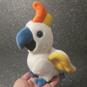 Puede incluir: Cacatúa de peluche blanco con pico azul, cresta amarilla y naranja y patas azules.