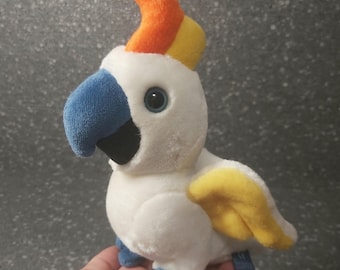 Könnte beinhalten: Weißer Plüschkakadu mit blauem Schnabel, gelber und orangefarbener Haube und blauen Füßen.