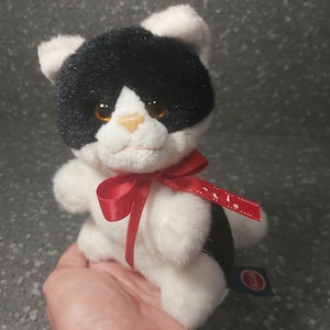 Op de afbeelding: Zwart-witte pluche katten speelgoed met een rode strik. De kat heeft oranje ogen en een roze neus. Het speelgoed is ongeveer 25 cm hoog.