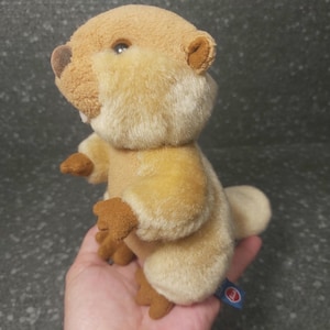 Op de afbeelding: Een pluche bever met een lichtbruin en bruin kleurenschema. Het knuffeldier heeft kleine bruine poten, oren en een staart. De bever heeft een vriendelijke uitdrukking met zichtbare tanden en een klein blauw label.
