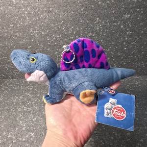 Puede incluir: Un juguete de dinosaurio de peluche azul con una vela morada y azul con lunares. El juguete tiene un llavero adjunto y una etiqueta que dice "Trudi". El dinosaurio tiene ojos amarillos y pies naranjas.