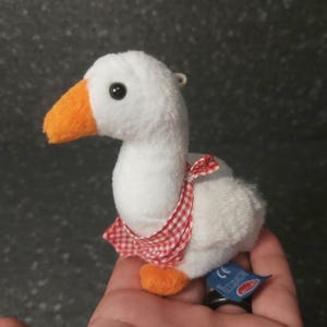 Puede incluir: Un peluche de ganso blanco con pico y patas naranjas, y un babero a cuadros rojos y blancos. El juguete tiene un pequeño lazo de metal para colgar y una etiqueta azul.