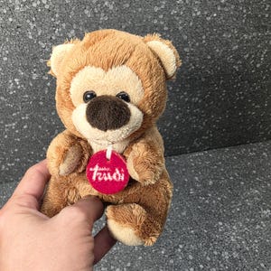 Op de afbeelding: Een kleine pluche teddybeer met lichtbruine vacht, een crèmekleurig gezicht en een donkerbruine neus. De beer heeft een rond, rood label om zijn nek hangen met de witte tekst Trudi erop gedrukt. Het speelgoed heeft twee kleine oren en pootjes.