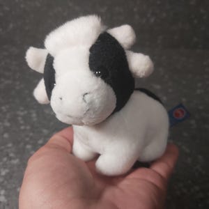 Op de afbeelding: Een klein, wit en zwart pluche koespeelgoed. De koe heeft een zwarte vlek op zijn gezicht en een witte vlek op zijn kop. Het speelgoed zit op de hand van een persoon.