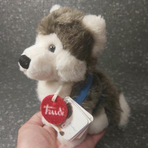 Op de afbeelding: Een pluche husky hondenspeelgoed met bruin en wit bont, een blauwe halsband en een rood label met het woord "Trudi" erop.