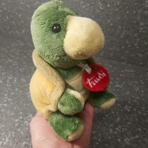 Può includere: Un peluche a forma di tartaruga con testa e zampe verdi, guscio giallo e muso color crema. Il giocattolo ha un'etichetta rossa con testo bianco. La tartaruga è morbida e coccolosa.